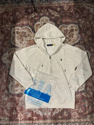 Grå hoodie från Polo Ralph Lauren - Snygg ljusgrå hoodie från Polo Ralph Lauren med dragkedja framtill och klassisk huva med dragsko. Broderad logga i marinblått på bröstet och ribbade muddar vid ärmar och midja. Tillverkad i mjuk bomullsblandning för extra komfort.