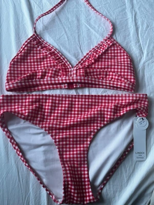 Rutig röd bikini aldrig använd - Snygg bikini från Lindex i rödvitt rutigt mönster. Överdelen har halterneck och lätt rynkade detaljer framtill. Bikinitrosan har klassisk passform och är helfodrad. Lapp kvar och aldrig använd