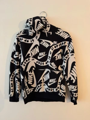 Nike hoodie med svartvitt tryck - Nike hoodie i svart med stort vitt all-over tryck av loggor och texten 'Just do it'. Klassisk huva och magficka framtill. Mjuk och bekväm, perfekt för en streetwear-look. Ribbade muddar vid ärmar och nederkant.