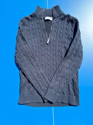 Select homme; Mörkblå kabelstickad halvzip tröja - Snygg mörkblå kabelstickad tröja med halvzip och hög krage. Tröjan har ribbade muddar vid ärmslut och nederkant, samt ett klassiskt kabelmönster över hela plagget. Perfekt för dig som gillar en stilren och tidlös look.