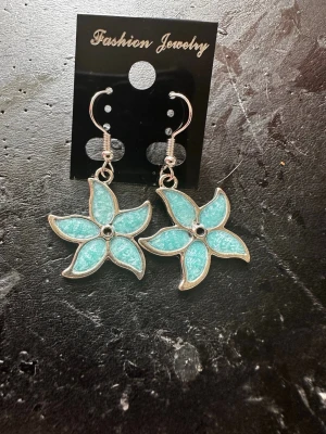 Turkosa blomformade örhängen - Säljer ett par snygga örhängen i form av blommor med fem blad. De är turkosa med silverfärgade kanter och har en glittrig finish. Perfekta för dig som vill ha något unikt och färgglatt till din stil.