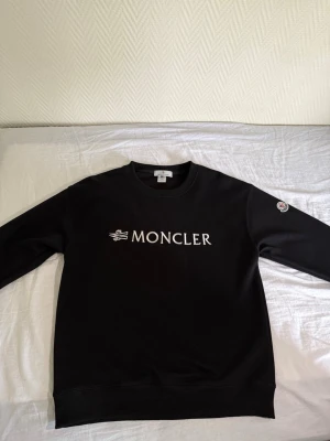 Svart Moncler sweatshirt - Snygg svart sweatshirt från Moncler med vit broderad logga på bröstet och klassisk Moncler-patch på ärmen. Tröjan har rund halsringning, ribbade muddar och är tillverkad i mjukt material för en clean och stilren look.