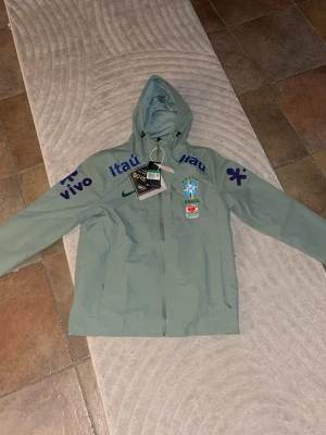 Nike Brasil vindjacka med huva - Ljusgrön vindjacka från Nike med huva och dragkedja. Jackan har Brasil-logga, flera sponsortryck i blått och grönt samt GORE-TEX-märke. Perfekt för dig som gillar sportig stil och vill sticka ut med landslagskänsla.