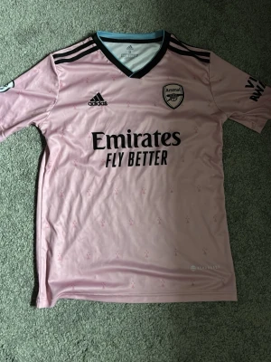 Arsenal rosa matchtröja Adidas S - Snygg rosa Arsenal matchtröja från Adidas med svarta detaljer och klubbmärke på bröstet. Tröjan har V-ringning med svart och ljusblå kant, samt tryckta loggor och sponsortexten 'Emirates Fly Better' på framsidan. Tillverkad i lätt och ventilerande material.