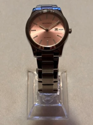 Sekonda klocka med rosa urtavla - Stilren klocka från Sekonda med metallarmband i silver och en snygg rosa urtavla. Klockan har datumvisning och klassisk rund form. Perfekt accessoar för dig som vill ha något både elegant och enkelt.