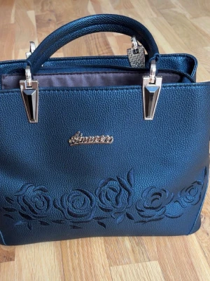 Svart handväska med rosdetaljer - Stilren svart handväska från Aimee i konstläder med präglade rosor framtill och guldiga metalldetaljer. Väskan har två handtag, flera innerfack och dragkedja. Perfekt för dig som gillar blommiga detaljer och vill ha en praktisk men snygg väska.💕