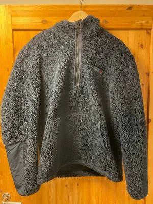Svart teddytröja från barbour  - Mysig svart teddytröja från Lundhags med halv dragkedja framtill och huva. Tröjan har stora fickor och förstärkta partier på armbågarna. Perfekt för kalla dagar och riktigt skön att bära. Materialet är fluffigt och värmande.