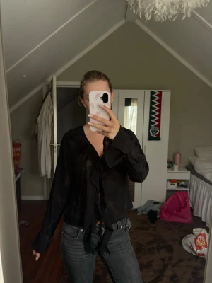 Svart transparent blus med rosett - Snygg svart blus i transparent material med diskret mönster. Blusen har långa ärmar och en dekorativ rosett framtill vid halsen. Perfekt för dig som gillar en trendig och lite mystisk look. Passar till både jeans och kjol.