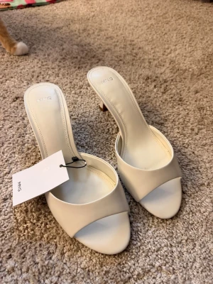 Vita klackar från Mango - Stilrena vita slip-in pumps från Mango med öppen tå och låg klack. Skorna har en bred rem över foten och är tillverkade i ett slätt material som ger en clean look. Perfekta för att lyfta vilken outfit som helst.