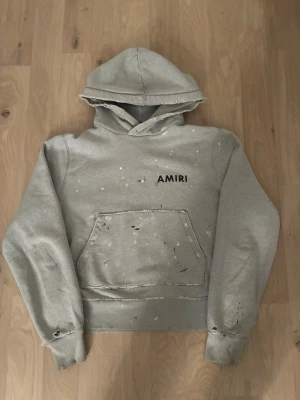 Grå hoodie från Amiri med tryck - Grå hoodie från Amiri med svart logotyptryck fram och bak. Hoodien har en stor magficka, huva och slitna detaljer för en edgy look. Tillverkad i mjukt material som känns skönt mot huden. Perfekt för dig som gillar streetwear och vill sticka ut.