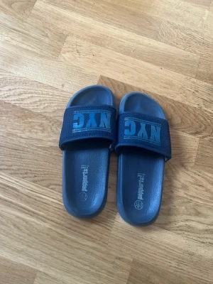 Mörkblå NYC badtofflor - Säljer ett par mörkblå badtofflor från Pepperts! med bred rem där det står 'NYC' i turkos text. Tofflorna har en klassisk slip-in modell och är tillverkade i syntetmaterial, perfekta för stranden eller hemmahäng. Perfekt för någon som behöver ett par extra tofflor om man ska simma. Möts upp helst i Malmö och Tar Swish, men kan fraktas spårbart! Pris kan diskuteras!