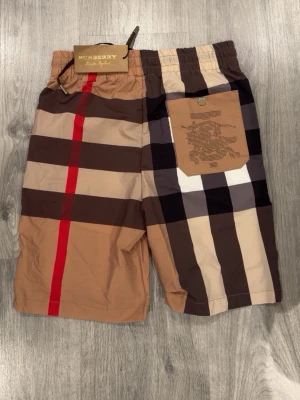 Rutig shorts från Burberry - Säljer ett par helt nya klassiska rutig shorts från Burberry med beige, bruna, svarta och röda toner. Shortsen har elastisk midja med snörning och en bakficka med Burberry-logga. Tillverkade i ett lätt syntetmaterial, perfekta för sommaren.