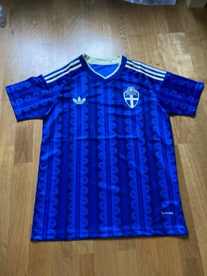 Blå Sverige Adidas matchtröja - Säljer en blå matchtröja från Adidas med Sveriges landslagsemblem på bröstet och vita Adidas-ränder på axlarna. Tröjan har ett unikt mönster i olika blå nyanser, V-ringning och korta ärmar. Materialet är lätt och andas, perfekt för träning eller match.
