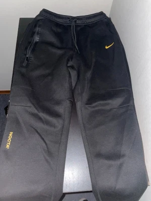 Svarta NOCTA mjukisbyxor från Nike - Säljer ett par svarta mjukisbyxor från Nike NOCTA med gul logga och detaljer. Byxorna har dragsko i midjan, fickor med dragkedja och broderade NOCTA- och Nike-detaljer. Perfekta för chill dagar och streetwear-stil.