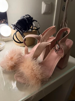 Beiga sandaler med fluffig detalj - Säljer ett par beiga sandaler med hög blockklack och justerbart spänne. Framtill har de en fluffig, fjäderliknande detalj som ger en unik och trendig look. Materialet ser ut att vara mockaimitation och innersulan är mjukt rosa. Perfekta för dig som vill sticka ut.