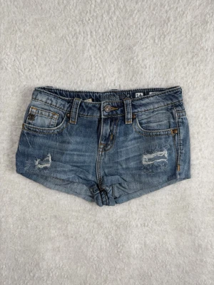 Miss me Blå denimshorts med slitningar - Säljer ett par blå denimshorts med slitna detaljer och klassisk femficksdesign. Shortsen har bälteshällor, knapp och dragkedja framtill samt råa kanter nedtill. Perfekta för varma dagar och en avslappnad stil.