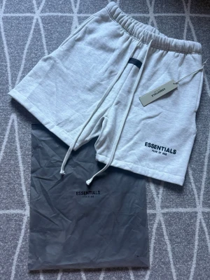 Grå shorts Essentials - Säljer ett par helt nya Snygga ljusgrå shorts från Essentials med svart logga på benet. De har elastisk midja med snörning och är tillverkade i mjuk bomull. Perfekta för nu för sommaren. Dustbag och etiketter tillkommer såklart.