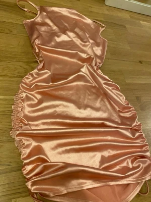 Rosa satin klänning med snörning - Snygg kort klänning i rosa satinmaterial med tunna axelband och snörning i sidorna. Klänningen har en figurnära passform och rynkade detaljer längs sidorna som ger en trendig look. Strl S och använt endast ett tillfälle!!