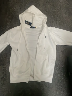 Vit hoodie från Polo Ralph Lauren - Snygg vit hoodie från Polo Ralph Lauren med dragkedja, två fickor framtill och klassisk huva med dragsko. Liten mörkblå logga broderad på bröstet. Tillverkad i mjukt material som känns skönt mot huden. Perfekt för en clean och stilren look.