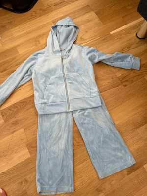Ljusblått velourset med huva - Mysigt loungewear-set i ljusblå velour med ziptröja och huva samt matchande byxor. Tröjan har fickor framtill och dragkedja med ringdetalj. Perfekt för chill dagar hemma eller när du vill vara extra bekväm.