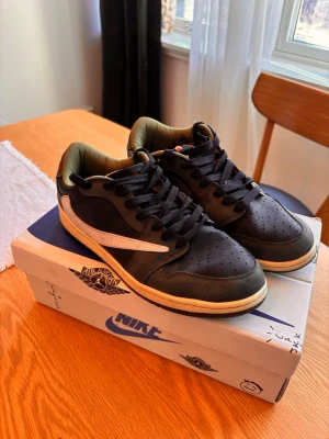 Nike Air Jordan 1 Low Travis Scott - Säljer ett par Nike Air Jordan 1 Low Travis Scott sneakers i svart och olivgrönt läder med den ikoniska omvända swoosh-loggan i vitt på utsidan och klassisk swoosh i oliv på insidan. Skorna har svarta skosnören, perforerad tåbox och beige mellansula. Röd broderad detalj på hälen.Är KOPIOR