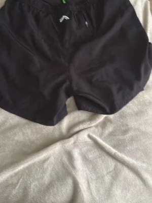 Svarta träningsshorts med snörning - Säljer ett par svarta träningsshorts med elastisk midja och snörning. Shortsen har två fickor med dragkedja på sidorna och är tillverkade i ett lätt syntetmaterial som passar perfekt för aktiva dagar.