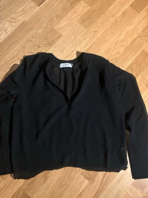 Svart blus från Selected Femme - Stilren svart blus från Selected Femme med v-ringning och långa ärmar. Blusen har en avslappnad passform och knappar vid ärmsluten. Perfekt att matcha med jeans eller kjol för en trendig look.