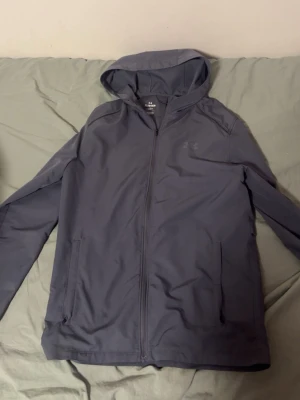 Mörkgrå jacka från Under Armour - Snygg mörkgrå hoodie från Under Armour med dragkedja framtill och huva. Tillverkad i ett sportigt, lätt material som passar perfekt till träning eller chill. Har två fickor och diskret logga på bröstet. Stilrena vita detaljer på axlarna ger en sportig look.