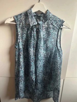 Ärmlös blus med blått mönster - Snygg ärmlös blus i tunnt, lätt material med blått och vitt abstrakt mönster. Blusen har en knytkrage framtill som ger en trendig touch. Perfekt att matcha med jeans eller kjol för en cool look.