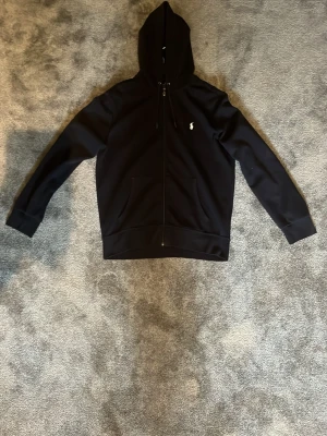 Svart hoodie från Polo Ralph Lauren - Klassisk marinblå hoodie med dragkedja från Polo Ralph Lauren. Har huva med dragsko, två fickor framtill och den ikoniska loggan broderad på bröstet. Perfekt för en avslappnad och stilren look.