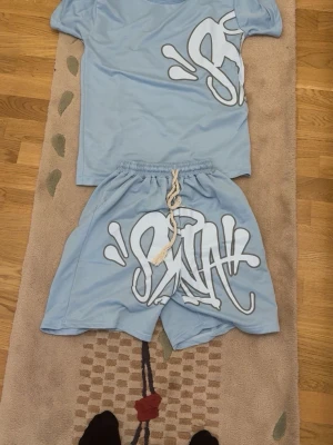 Ljusblå shorts med graffiti-tryck - Säljer ett par ljusblå shorts med stort vitt graffiti-inspirerat tryck framtill och snörning i midjan. Perfekta för dig som gillar streetwear och vill sticka ut med en unik stil. Matchande t-shirt finns också om du vill köra hela looken.