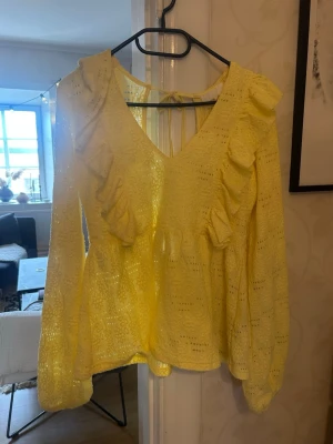 Gul blus med volanger och öppen rygg - Snygg gul blus med volangdetaljer framtill och längs ärmarna. Blusen har djup v-ringning, öppen rygg med knytband och är gjord i ett genombrutet spetsliknande material. Perfekt för dig som vill sticka ut med färg och detaljer.