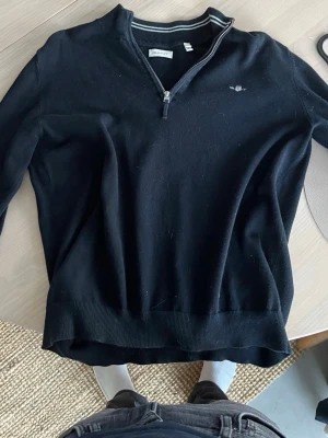 Svart halvzip-tröja från Gant - Svart långärmad tröja från Gant med halv dragkedja och broderad logga på bröstet. Tröjan har ribbade muddar och krage med randig detalj på insidan. Perfekt för en stilren och avslappnad look.