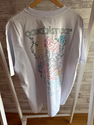 Vit Casablanca t-shirt med grafiskt tryck - Säljer en vit t-shirt från Casablanca med ett stort grafiskt tryck på ryggen i flera färger och texten 'casablanca' upptill. T-shirten har klassisk passform och korta ärmar. Perfekt för dig som gillar unika prints och stilrena plagg.