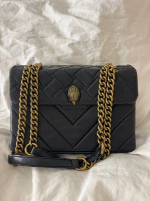 Kensington väska - Kurt Geiger London - Svart Kensington väska från Kurt Geiger med guldfärgad kedjerem och ikonisk örndetalj på locket.                                                                        Jag är fullt medveten om att väskan är använd och har en del slitage, vilket syns på bilderna. Det är viktigt att påpeka att väskan inte har några hål, revor eller andra skador i materialet – det handlar endast om att lädret nötts på vissa ställen efter användning. Har fler bilder om det önskas