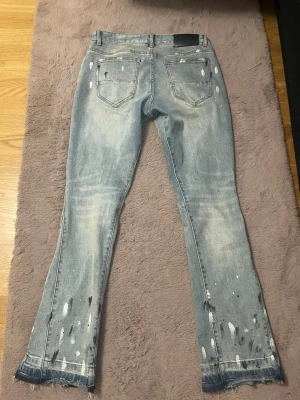 Ljusblå Lorenzo jeans med slitningar - Säljer ett par ljusblå jeans från Lorenzo med coola slitningar, färgstänk och råa kanter vid bensluten. Jeansen har två bakfickor och en tvättad look som ger en trendig och avslappnad stil. Perfekta för dig som gillar streetwear och unika detaljer.