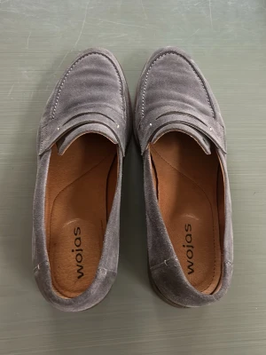 Grå mockaloafers från Wojas - Snygga loafers i grå mocka från Wojas med klassisk pennyloafers-design. Skorna har en låg klack i trälook och mjuk innersula för extra komfort. Perfekta för dig som gillar stilrena och tidlösa skor. pris går att diskutera