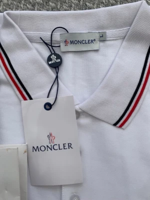 Vit pikétröja från Moncler - Säljer en klassisk vit pikétröja från Moncler med krage och knappar framtill. Tröjan har snygga röda och svarta ränder på kragen samt Moncler-logga broderad på bröstet. Tillverkad i bomull för en skön och luftig känsla.