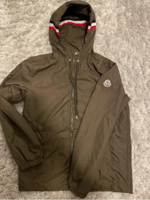 Olivgrön Moncler vindjacka med huva - Snygg olivgrön vindjacka från Moncler med dragkedja, justerbar huva och elastiska muddar. Jackan har Moncler-logga på ärmen och en cool randig detalj i rött, vitt och blått på huvan. Tillverkad i lätt och tåligt material, perfekt för blåsiga dagar.