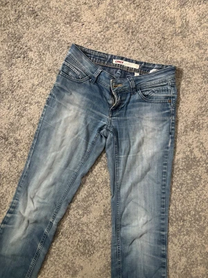 Ljusblå lågmidjade bootcut jeans från ONLY - Säljer ett par ljusblå bootcut jeans från ONLY med klassisk femficksdesign och slitna detaljer framtill. Jeansen har normal midja och dragkedja med knapp. Perfekta för en avslappnad och trendig look.