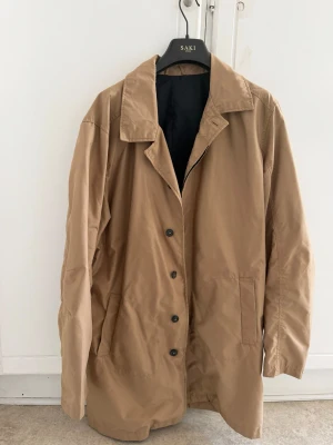 Beige rock från Jack & Jones - Stilren beige rock från Jack & Jones med klassisk krage och knäppning framtill. Rocken har två stora fickor på sidorna och en innerficka med dragkedja. Fodrad insida i svart tyg och detaljer i samma beige ton. Perfekt för dig som vill ha en enkel men snygg look.