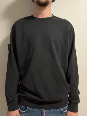 Svart sweatshirt från Stone Island - Svart långärmad sweatshirt från Stone Island med klassisk rund hals och ikonisk logotyp-patch på vänster ärm. Tröjan har en avslappnad passform och är tillverkad i mjuk bomull. Perfekt för en stilren och enkel look.