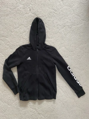 Svart hoodie från Adidas med tryck - Säljer en svart hoodie från Adidas med dragkedja, huva och fickor framtill. Adidas-logga på bröstet och stort vitt tryck längs ena ärmen. Tillverkad i mjuk bomullsblandning, perfekt för en sportig och avslappnad stil.