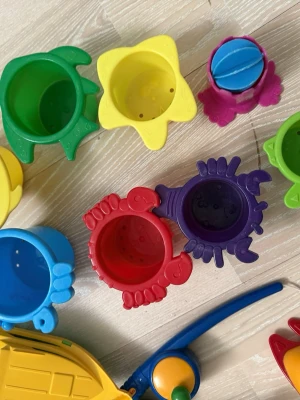 Färgglada badleksaker och stapelkoppar - Säljer ett set med färgglada badleksaker och stapelkoppar i plast, formade som djur och hinkar. Perfekt för lek i badet eller sandlådan. Inkluderar även en liten båt och en badanka. Alla delar är i olika färger som röd, blå, grön, gul, lila och orange.