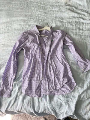 Lila rutig skjorta från Ralph Lauren - Snygg lila och vit rutig skjorta från Ralph Lauren med klassisk krage och grönt broderat logotyp på bröstet. Skjortan är i bomull med stretch och har långa ärmar samt knappar framtill.