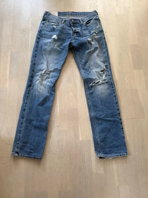 Blå slitna jeans från Hollister - Ett par blå jeans från Hollister med raka ben och tydliga slitningar och hål på framsidan. Jeansen har klassisk femficksdesign, knappgylf och en skinnpatch med logga bak i midjan. Perfekta för en avslappnad och trendig look.