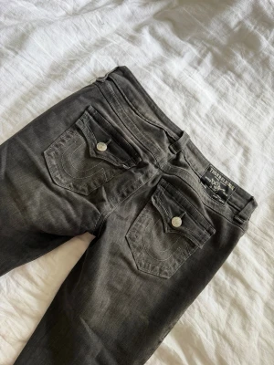 Svarta jeans från True Religion - Säljer ett par svarta jeans från True Religion med ikoniska ficklock och kontrastsömmar. Jeansen har raka ben, silverfärgade knappar och detaljerade bakfickor med logga. Perfekta för dig som gillar streetwear och vill ha en cool look.