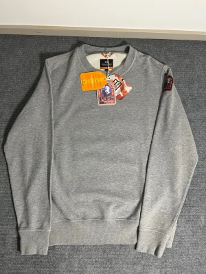 Parajumpers – Grey Sweatshirt with Yellow Back (L) - Brand new Parajumpers sweatshirt in stylish grey with a striking yellow back panel. Size L, length 68 cm, width 58 cm – fits comfortably with a premium feel. The back features a bold PARAJUMPERS print across the yellow section, making it an eye‑catching and unique design.  ✅ Condition: Unworn – completely untouched, in perfect condition 💯 Authenticity: 100% genuine, can be verified easily on certilogo.com 🧵 Material: Thick, warm, and high‑quality cotton blend – soft, durable, and comfortable 📦 S