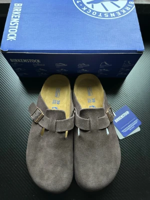 Grå mockatofflor från Birkenstock - Säljer ett par gråa Birkenstock tofflor i mocka med klassisk slip-in design och justerbart spänne i metall. Tofflorna har en mjuk innersula och präglad logga på sidan. Perfekta för chill dagar hemma eller när du vill ha det bekvämt.
