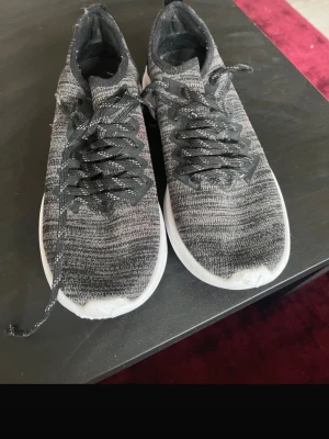Svarta stickade sneakers från Puma - Säljer ett par svarta och gråmelerade sneakers från Puma med stickad ovandel och vita sulor. Skorna har svarta snören med vita detaljer och stor PUMA-logga bak på hälen. Perfekta för en sportig och avslappnad stil.